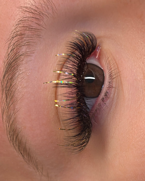 alt="Close-up of lash set with black volume fans and gold tinsel strands. Wispy textured lash look with added sparkle using glitter lash extensions."